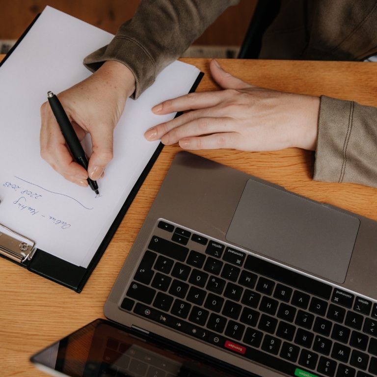 Man sieht einen Schreibtisch von oben. Darauf befindet sich ein Laptop und ein Schreibblock. Man sieht zwei Hände, wobei eine Hand mit einem Stift etwas notiert.