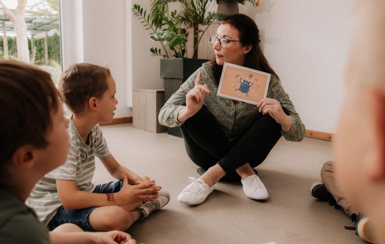 Evamaria Gerber sitzt mit Kindern auf dem Boden im Kreis. Sie hält eine Bildkarte in der Hand, hebt eine Hand und schaut ein Kind an.